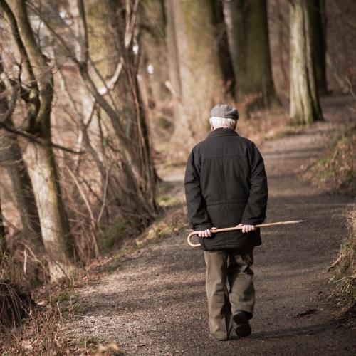 Ein älterer Herr geht alleine auf einem Waldweg spazieren. Das Bild zeigt ihn von hinten, wie er einen Spazierstock hinter seinem Rücken mit beiden Händen hält.